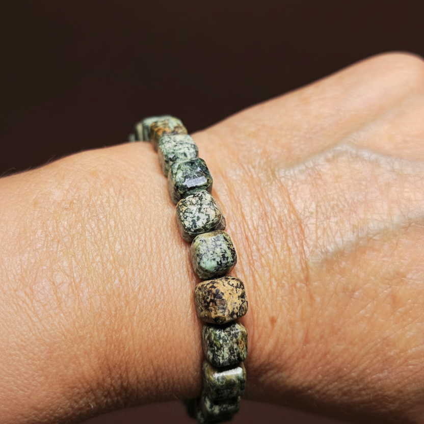A close-up of a person's wrist wearing a beaded bracelet made of rough, polished stone beads in earthy green, brown, and black tones.