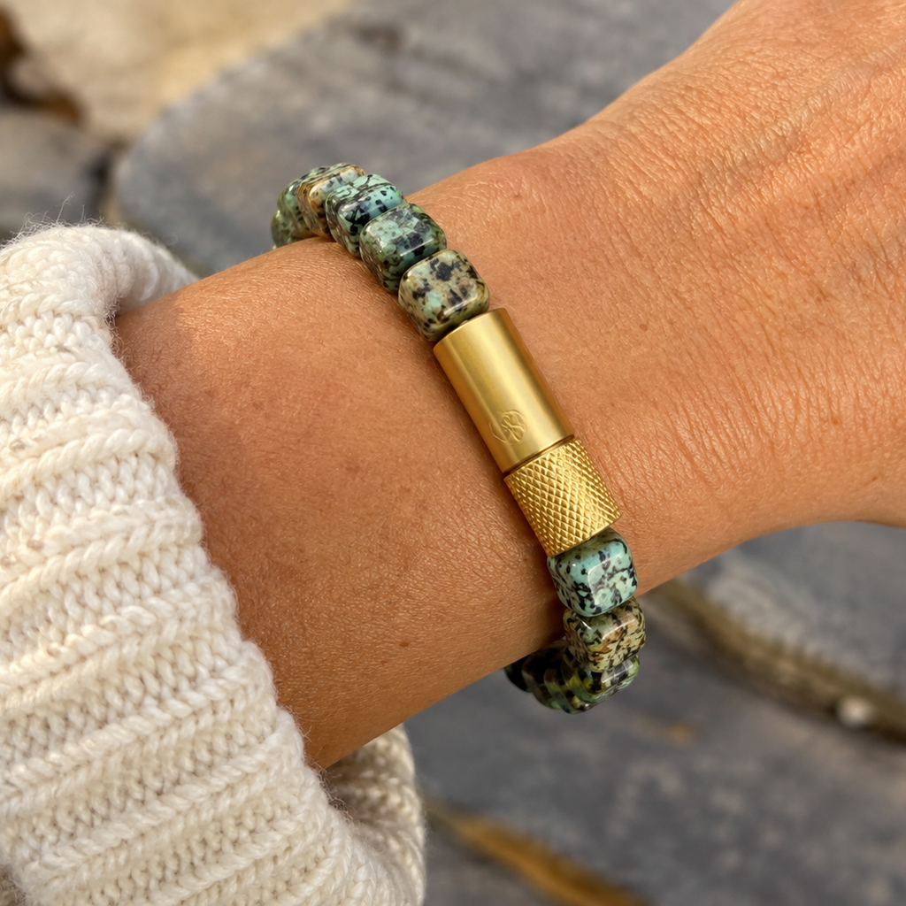 A close-up of a person's wrist wearing a beaded bracelet with turquoise-like green and black beads and a prominent gold-tone cylindrical clasp, with a cream knit sleeve visible.