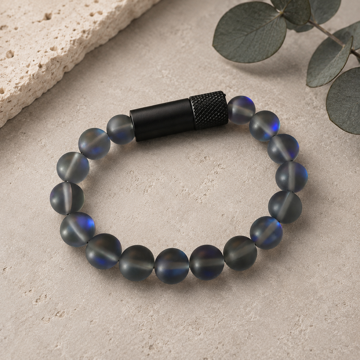 A beaded bracelet with matte gray-blue oval beads showing a subtle iridescent blue sheen, secured by a textured black cylindrical clasp, displayed on a light stone surface with decorative leaves.