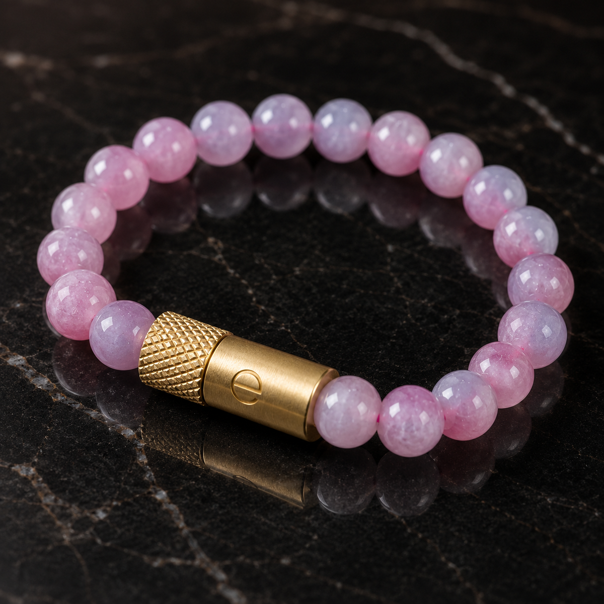 A beaded stretch bracelet made of round pink stone beads with a textured gold metal clasp, displayed on a dark marble surface.