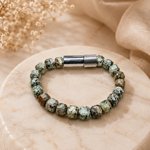 A beaded bracelet with green and white marbled stone beads and a textured silver clasp, displayed on a round beige marble tray with dried flowers and soft beige fabric in the background.