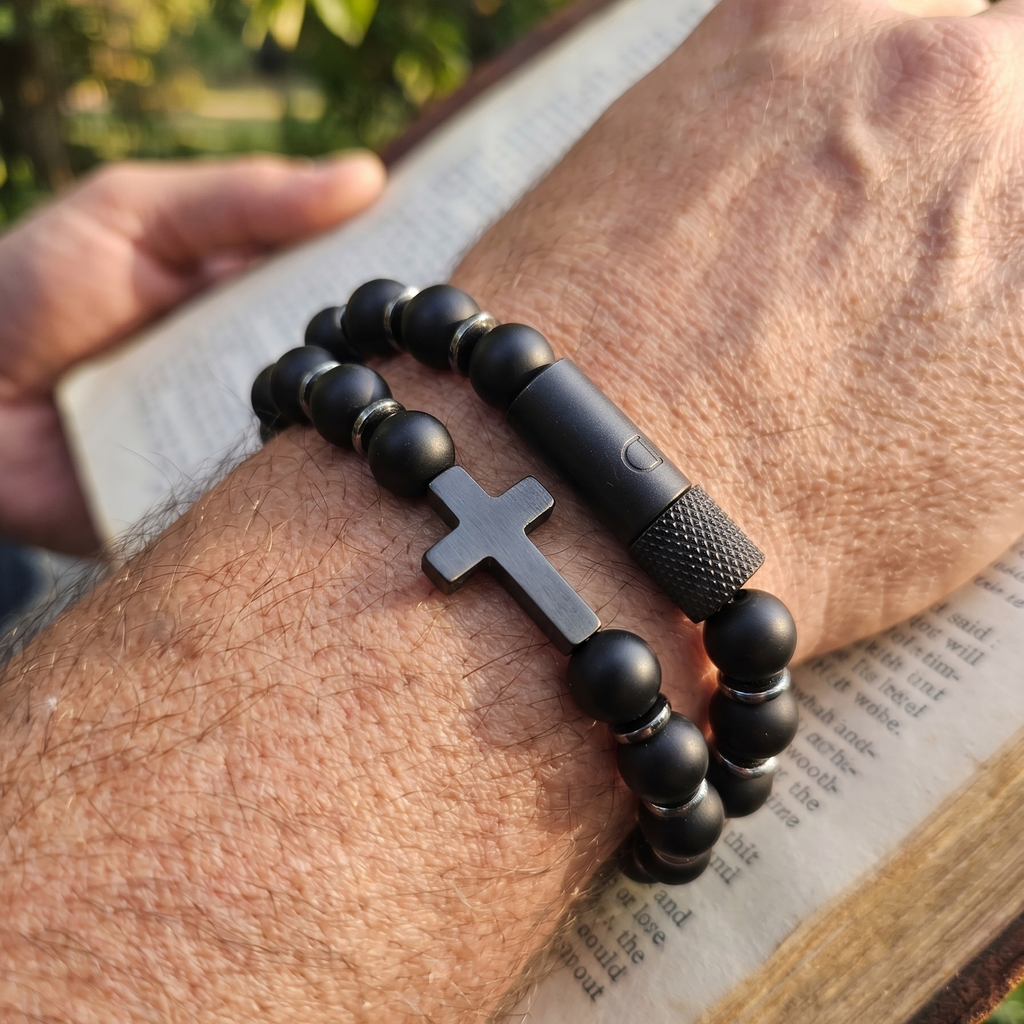 Black beaded bracelet with a cross pendant on a wrist, held over an open book outdoors.