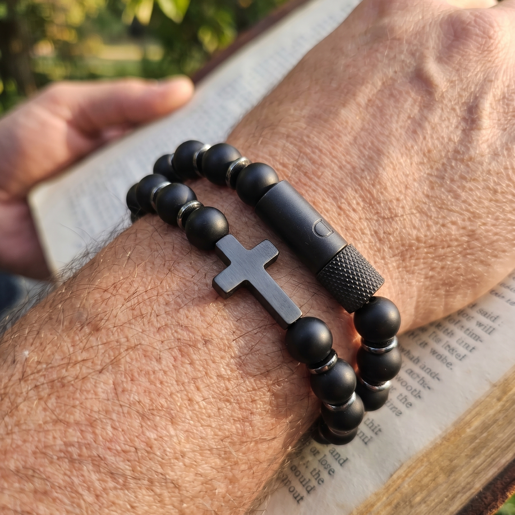 Black beaded bracelet with a cross pendant on a wrist, held over an open book outdoors.