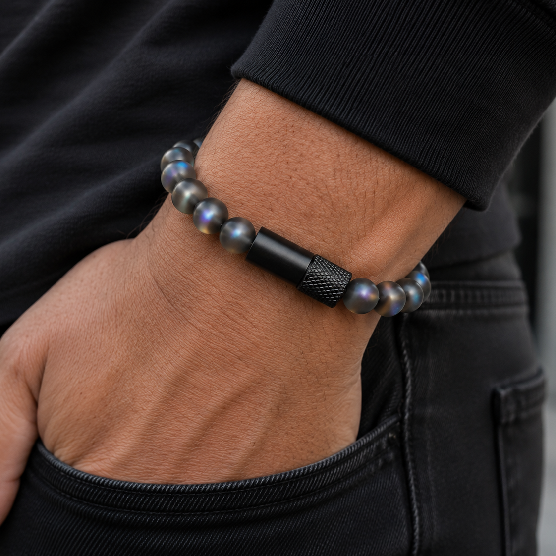 A close-up of a person's wrist wearing a beaded bracelet with dark gray iridescent beads, a black cylindrical accent, and a knurled black metal segment, styled with a black long-sleeve shirt and dark jeans.