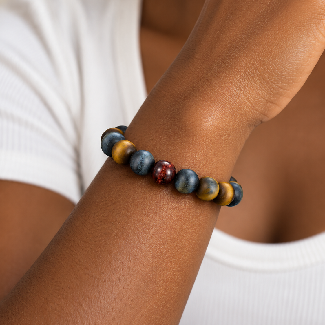 A close-up of a person's wrist wearing a beaded bracelet made of round blue-gray beads, one gold bead, and one reddish-brown bead, against a white ribbed top.